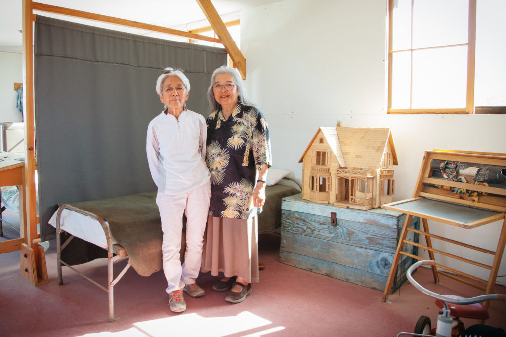 Former Manzanar child internees Irene Hadeishi, left, and her sister Marge Taniwaki, right, in a re-created children’s room. In order to make the re-creations of living spaces accessible, National Parks was required to make the living quarters much nicer than what was the reality for Irene and Marge and other families. They slept in cots separated only by sheets from other families with floors made of wood beams with space between them so the dust would come up constantly from the ground into their eyes and mouths. Photo by Joanne Kim