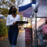 Young waitress serving drinks to customers in outdoor plastic tent wearing a mask. Waiter wearing protective face mask serving drinks at street cafe during pandemic.