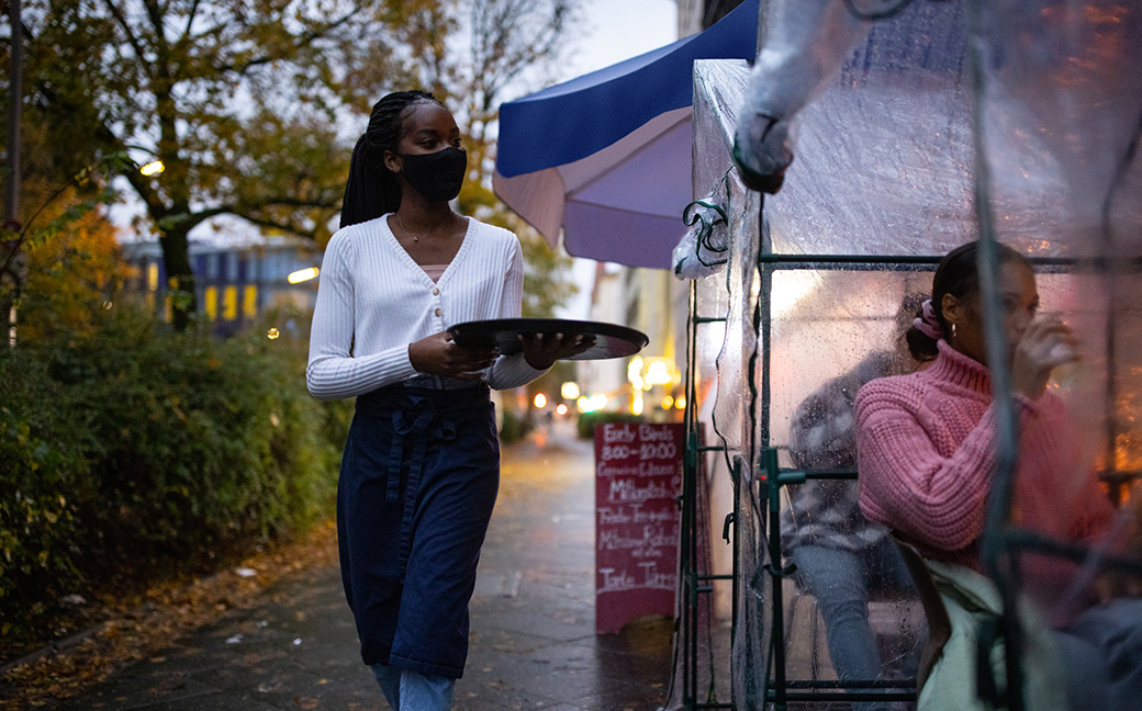 Young waitress serving drinks to customers in outdoor plastic tent wearing a mask. Waiter wearing protective face mask serving drinks at street cafe during pandemic.